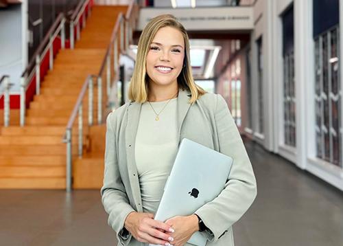 A young, professionally-dressed woman holding an Apple laptop while standing in front of you